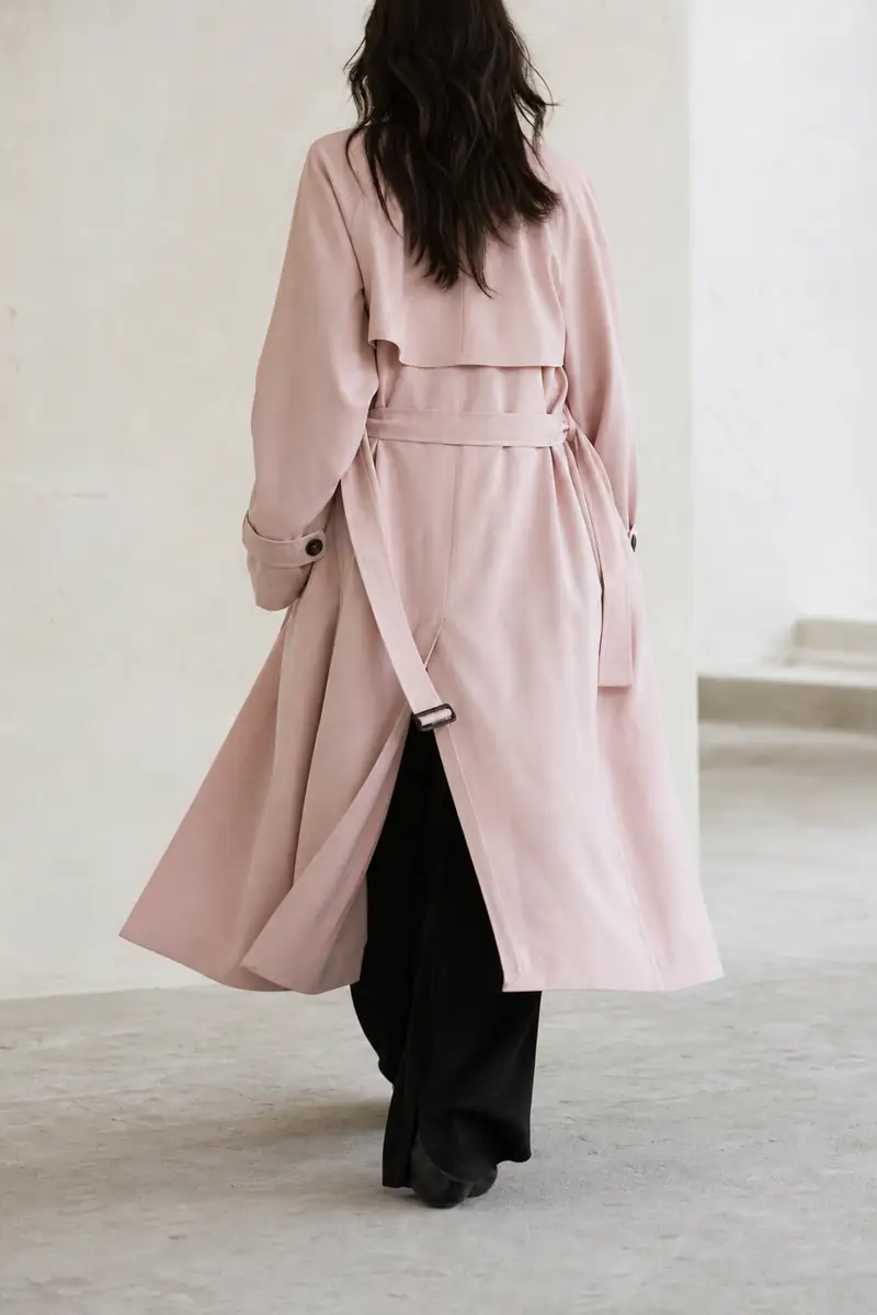 Donna di spalle con un lungo trench rosa cipria e pantaloni neri larghi